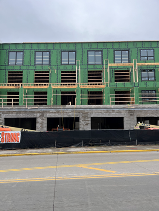 Building under construction with green sheathing, windows, and scaffolding. Black fencing in front.
