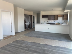 Empty open-concept apartment with wood-look flooring, white walls, and a partial kitchen with dark cabinets.