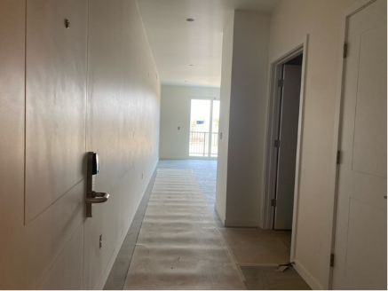 Hallway with white walls, a door, and a glimpse of a room with a balcony.