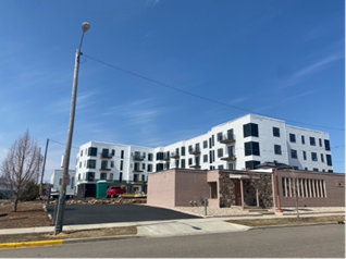 White apartment building under construction, blue sky. Concrete sidewalk, street lamp, and a small brown building in front.