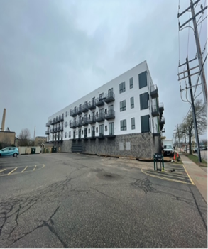 Modern white and stone apartment building with balconies, gray sky, parking lot in front.