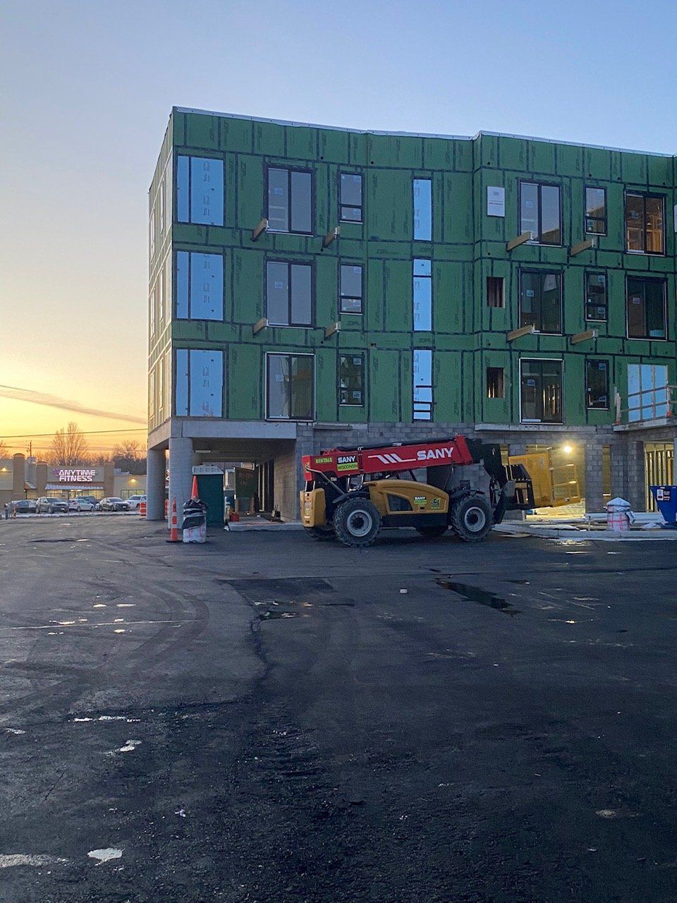 Building under construction with a yellow lift in front. Exterior walls are covered in green panels.
