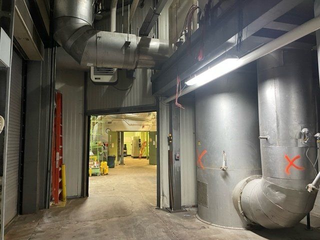 Hallway with large pipes, doorways, and bright lighting in a utility or industrial space.