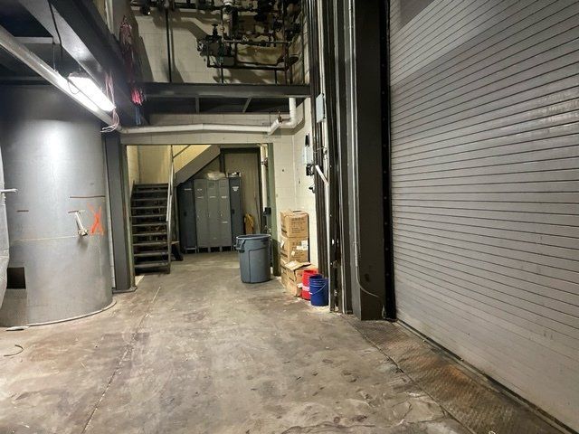 Industrial hallway with a roll-up door, metal pipes, stairs, and large cylindrical structure.