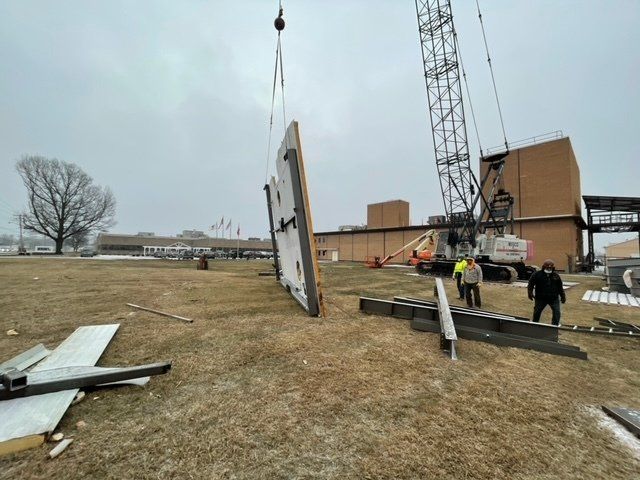 A crane lifts a large panel. Workers in a field near a building under overcast skies.