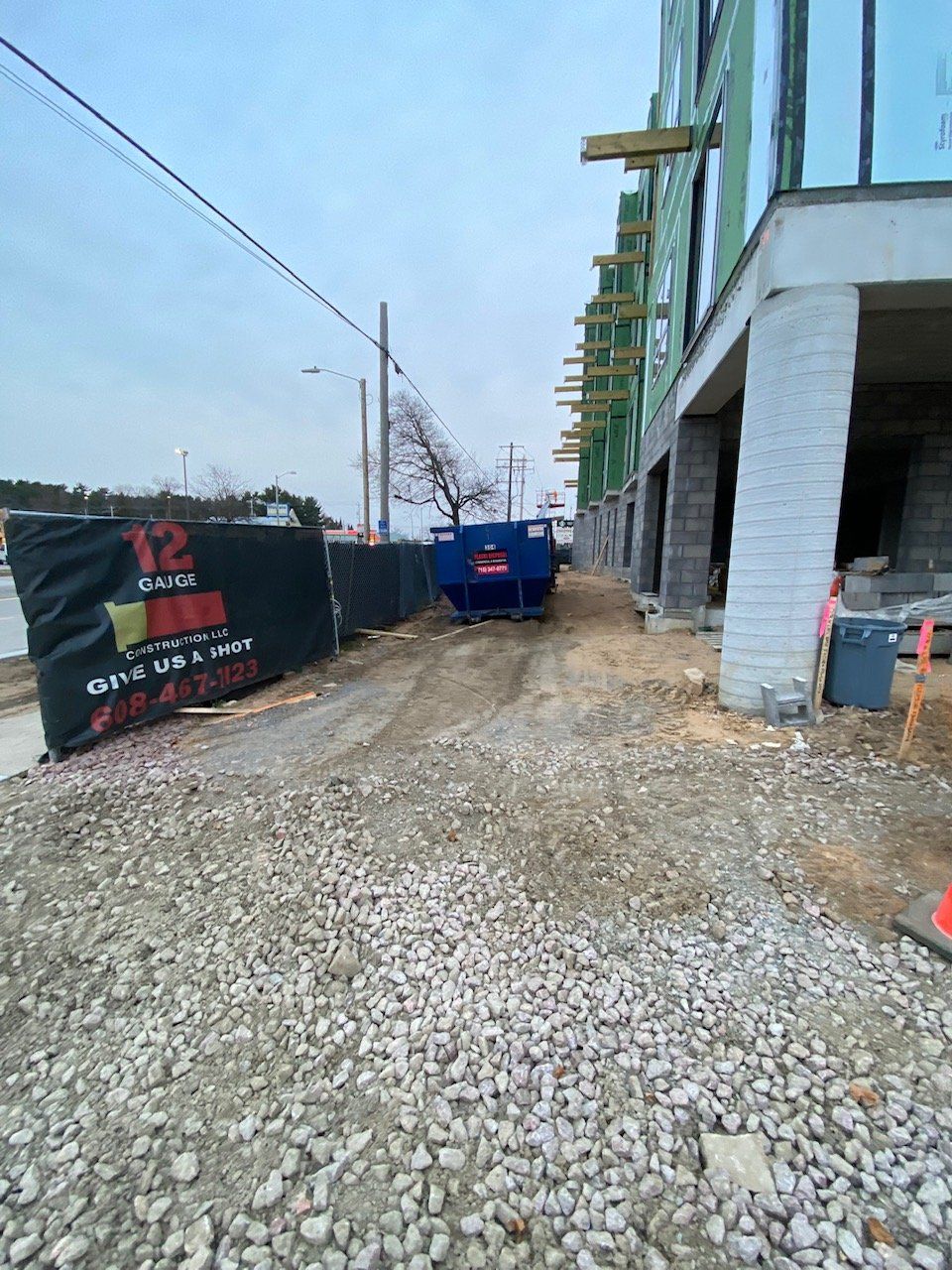 Construction site: Blue dumpster on gravel path between building and black fence.
