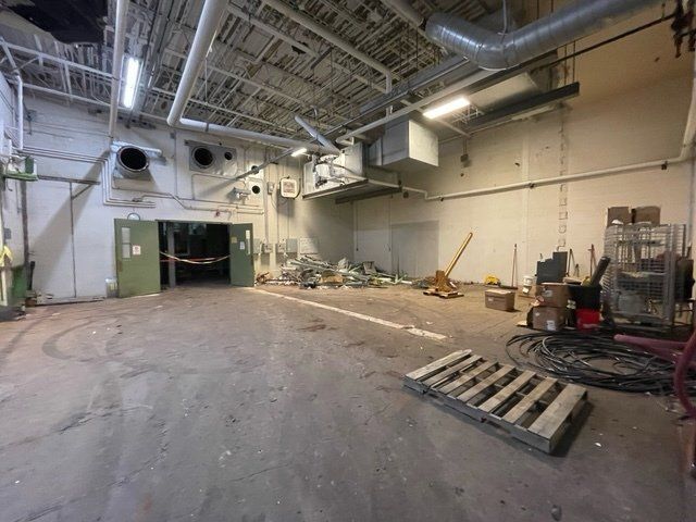 Interior view of a large, cluttered industrial space with concrete floor, air ducts, and construction debris.
