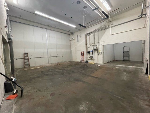 Empty industrial room with concrete floor, white walls, exposed pipes and overhead lighting.