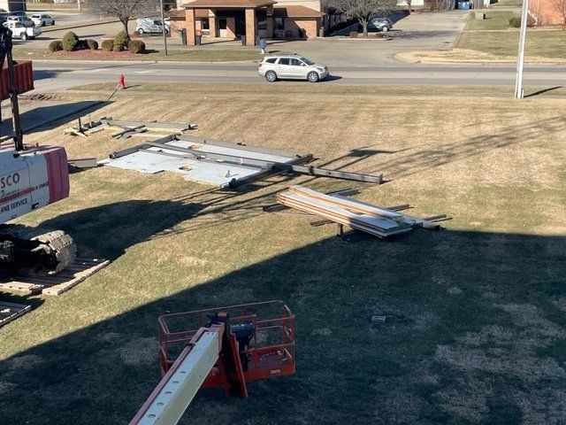 Deconstructed sign parts on the grass, a cherry picker, a road, and a building in the background.