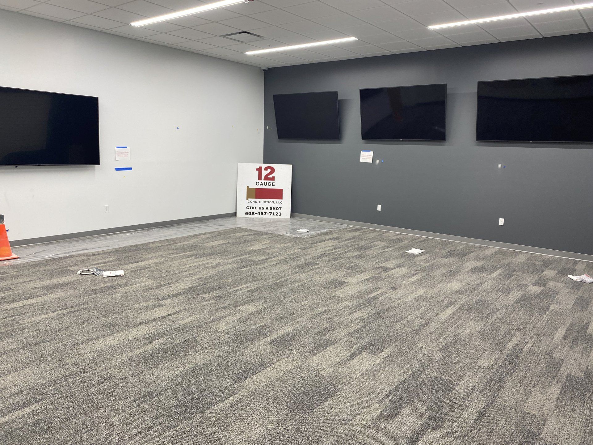 Empty room with grey carpet, white and gray walls, three wall-mounted TVs, and a sign on the floor.