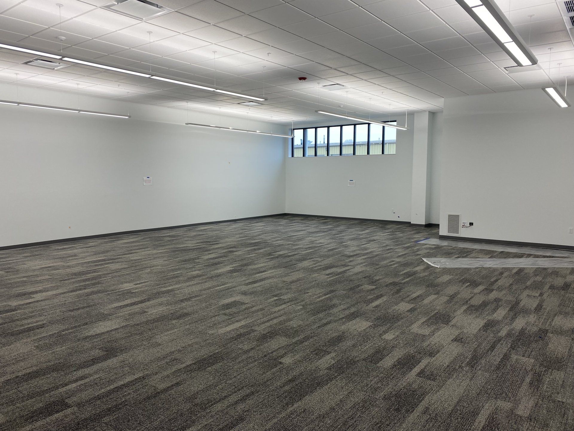 Empty office room with gray patterned carpet, white walls, and ceiling lights.