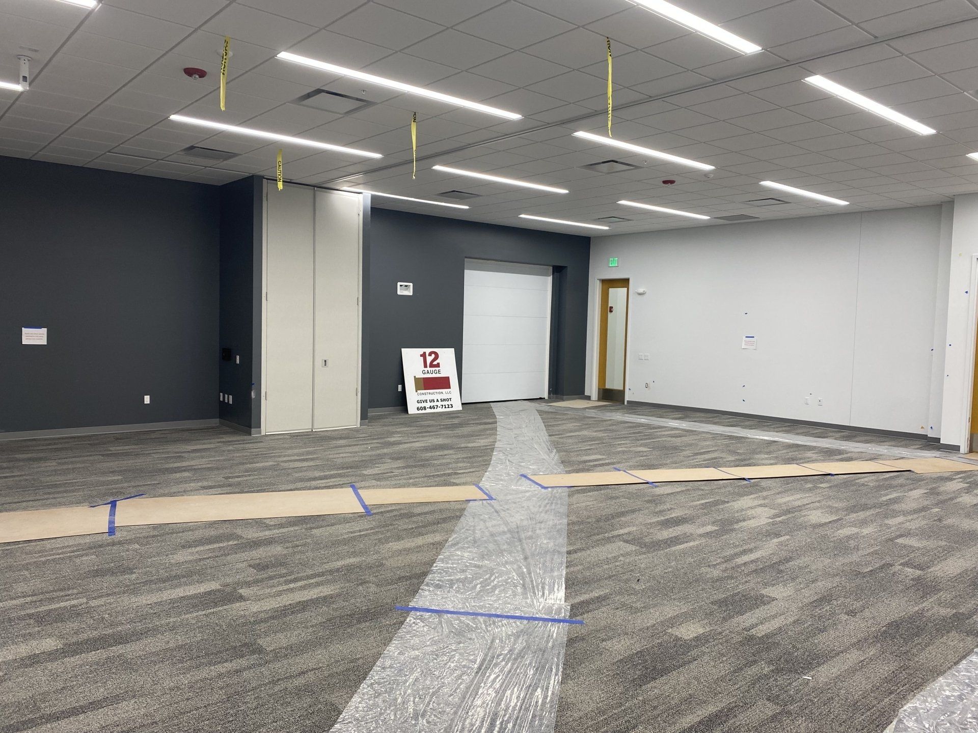 Empty room with gray carpet, gray and white walls, overhead lights, and a closed white door.