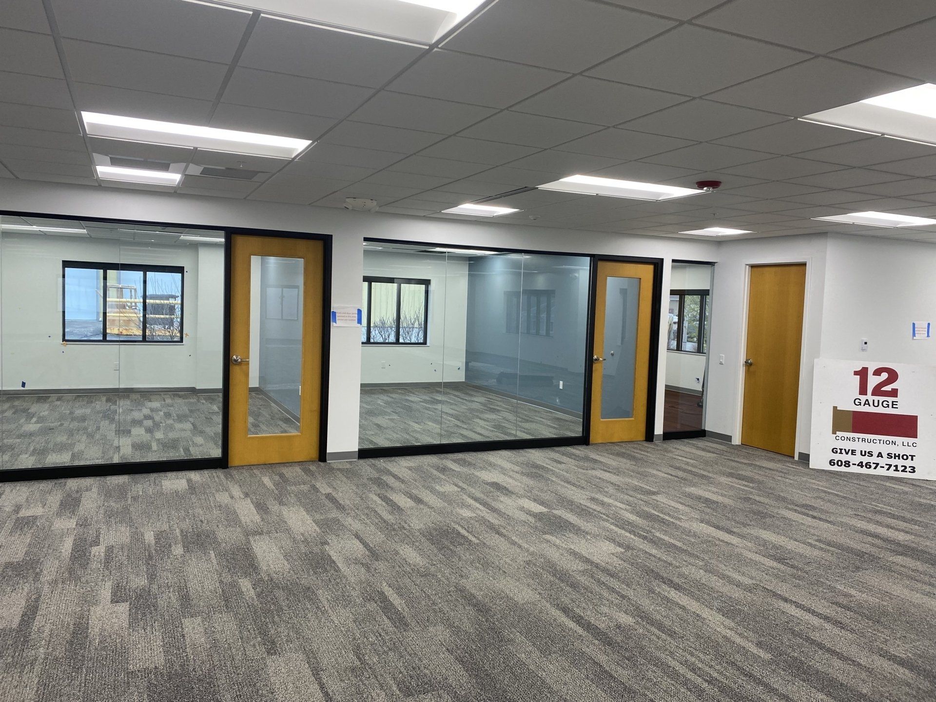 Empty office space with glass-walled rooms and carpeted floor. Doorways with wooden frames. Fluorescent ceiling lights.