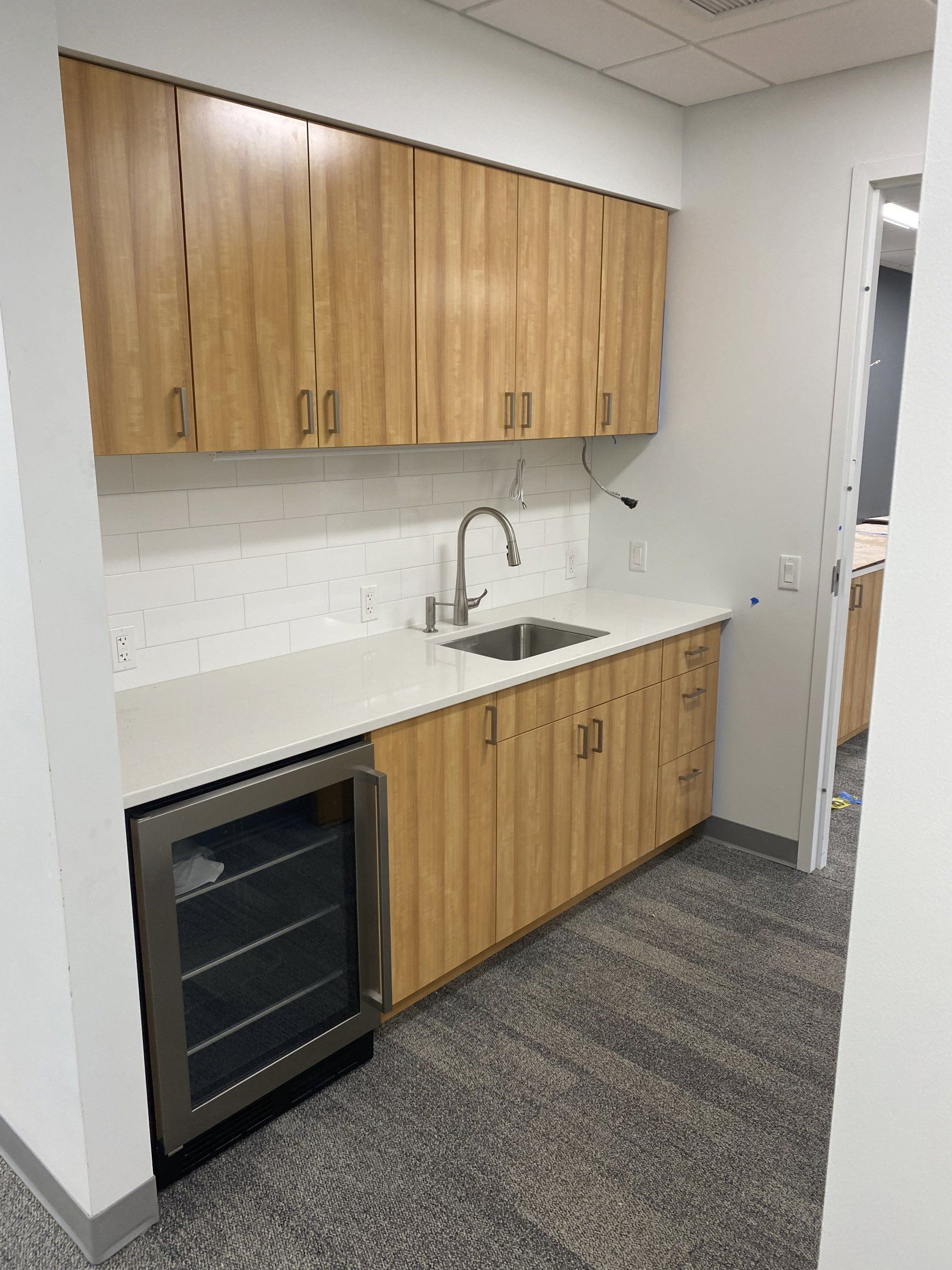Kitchenette with light wood cabinets, white countertop, stainless steel sink, and a beverage cooler.