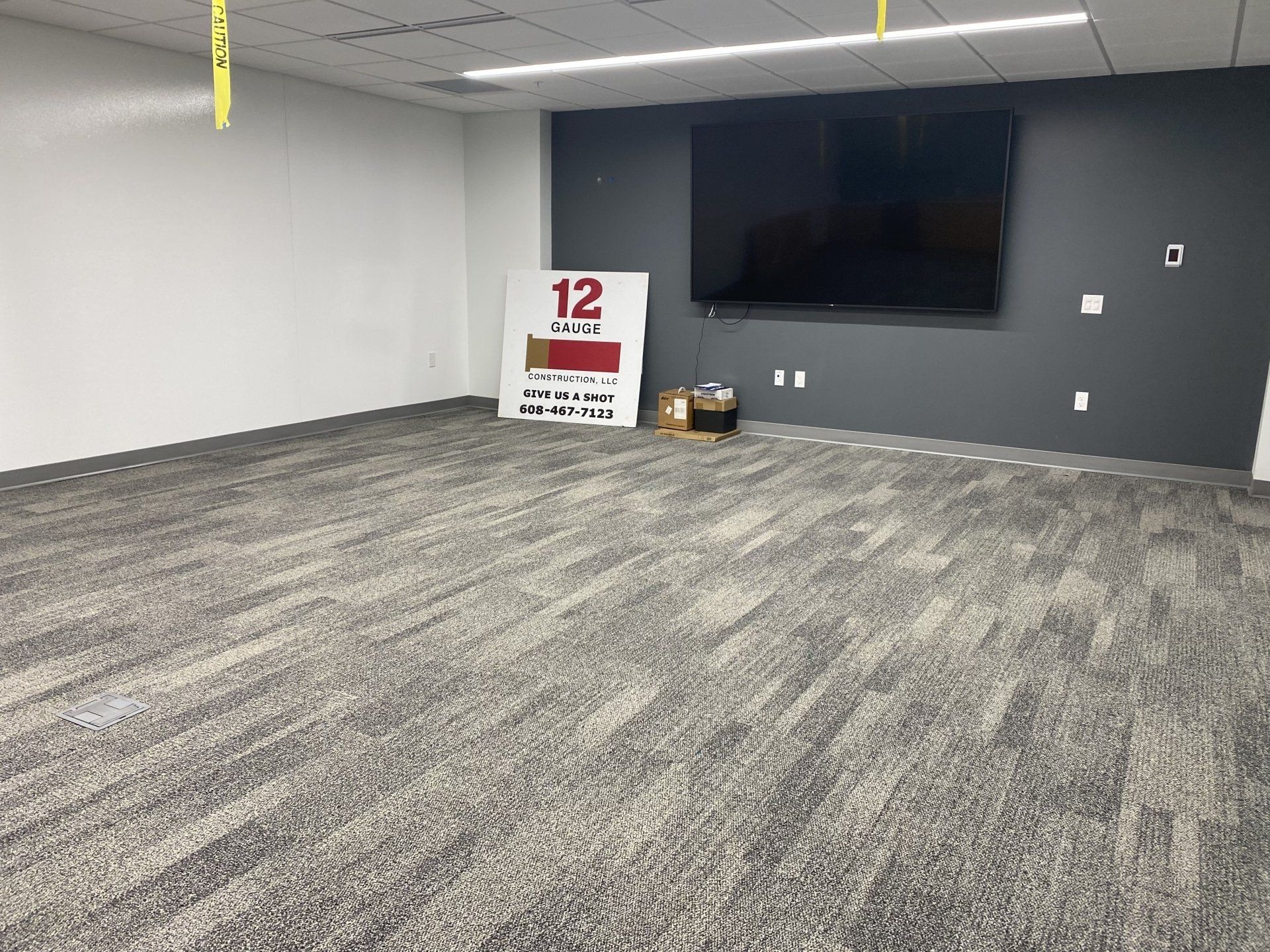 Empty room with gray patterned carpet, large TV on dark gray wall, white wall, and a sign.