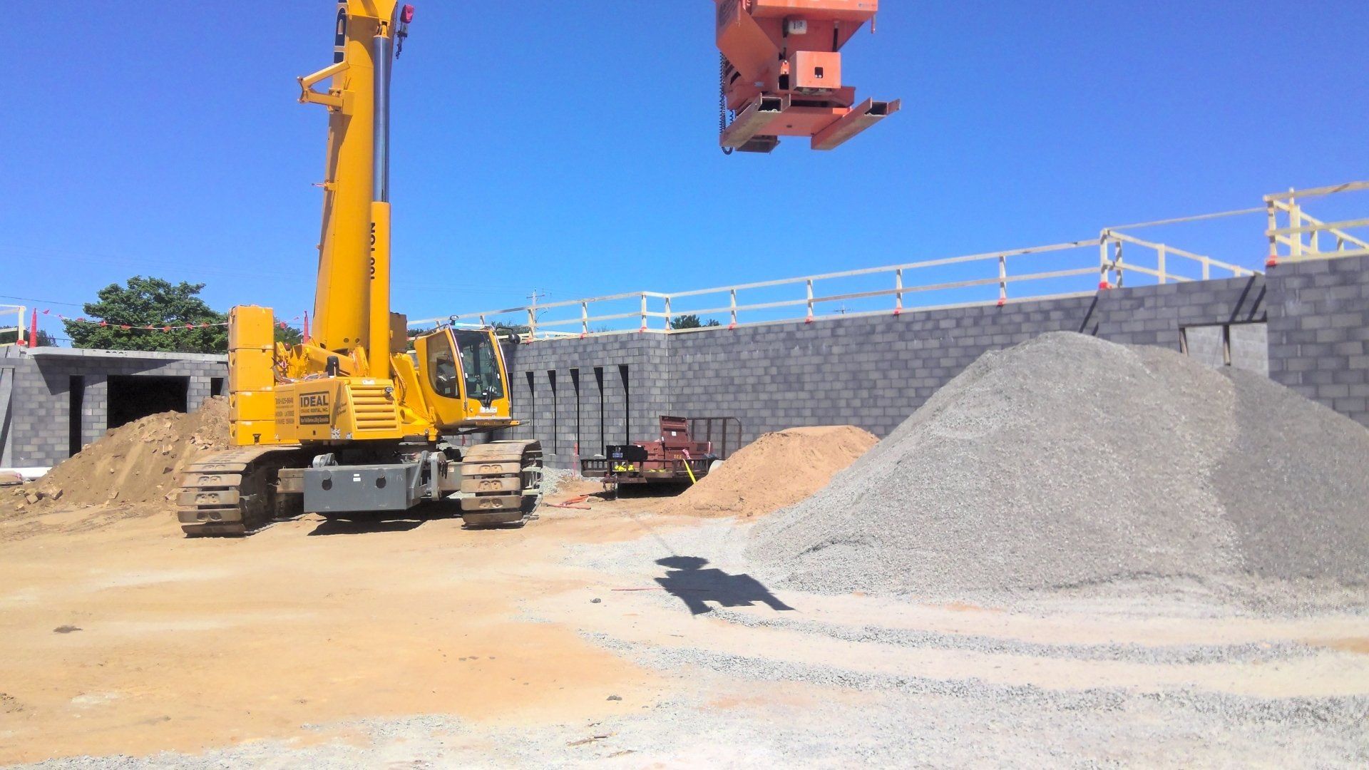 Construction site with yellow machinery and piles of gravel and sand. A concrete block wall is in the background.