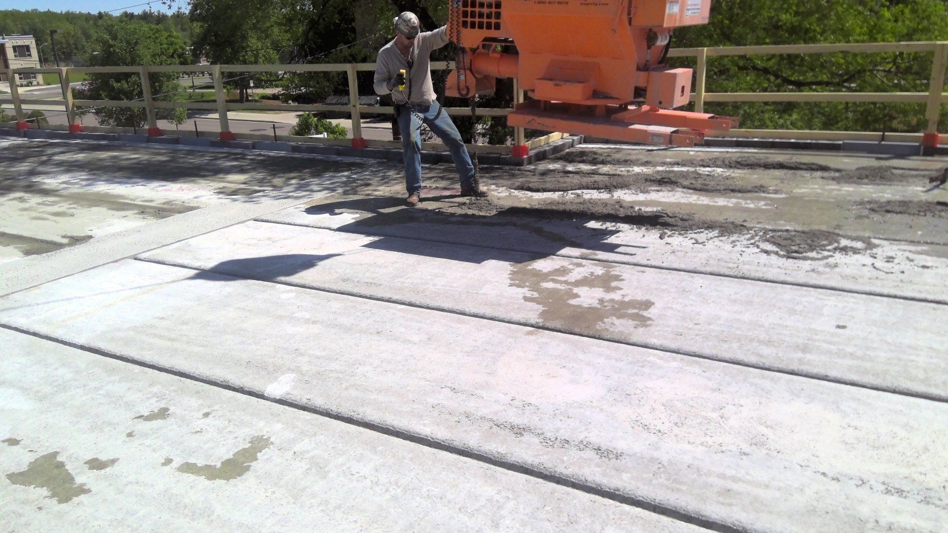 Man operating concrete pouring machine on bridge deck. Orange equipment, concrete surface, yellow safety rails.