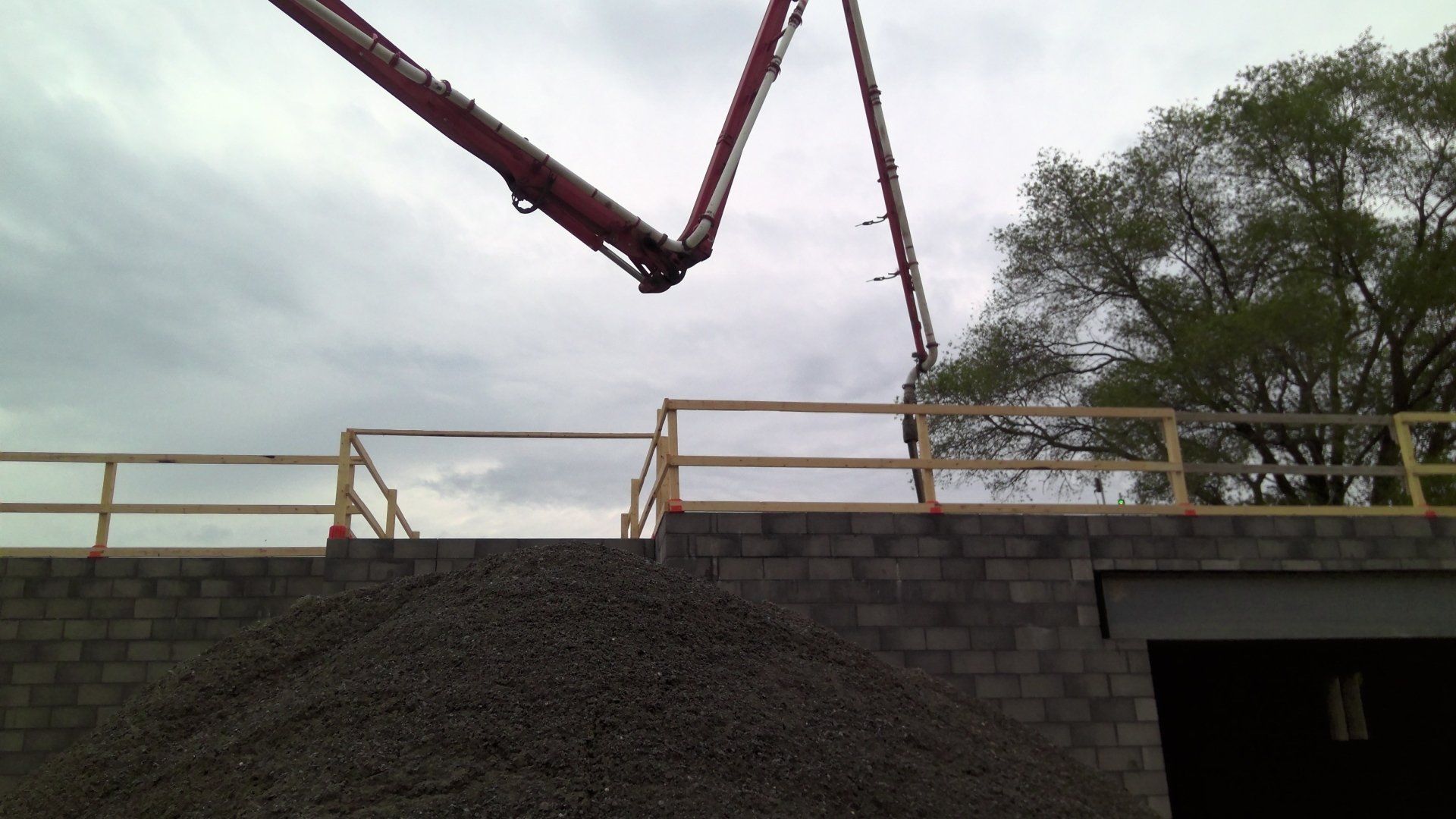 Concrete pump arm pouring concrete onto a construction site. Gray block wall, yellow railing, pile of rocks, cloudy sky.