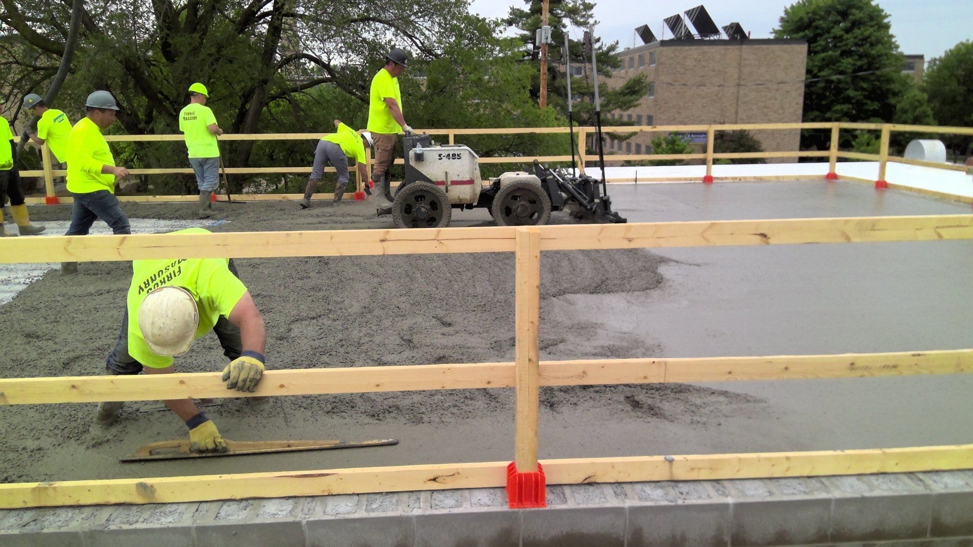 Construction workers in neon shirts pour and level concrete on a rooftop. Safety railing in foreground.