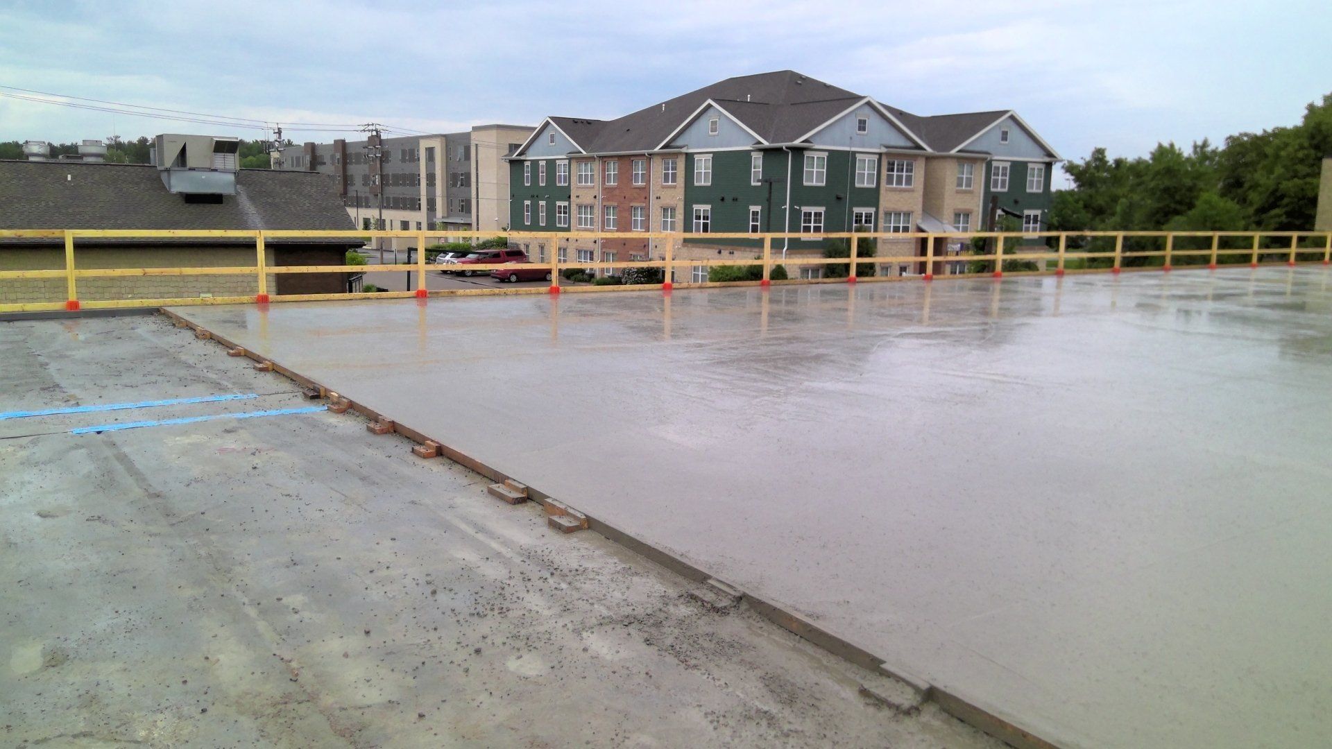 Wet concrete on a flat rooftop, with safety barriers in place. Apartment building in background. Cloudy sky.