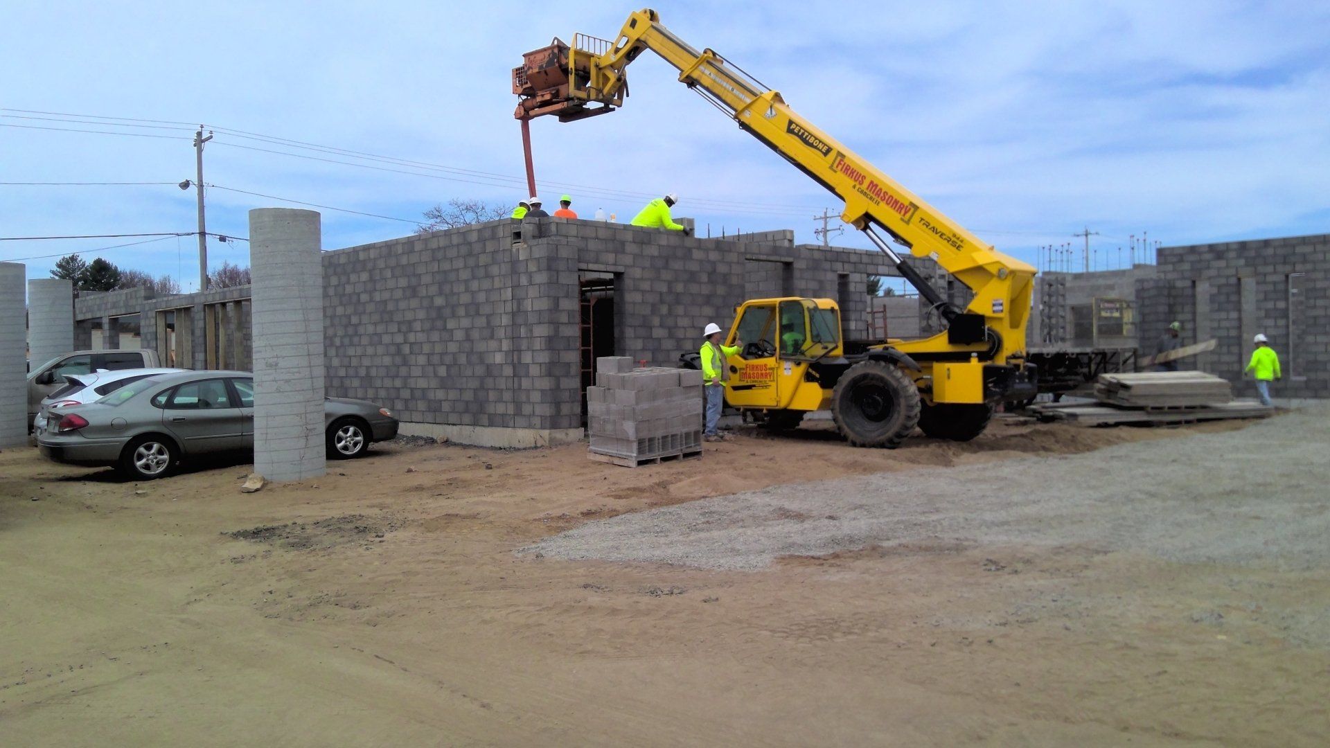 Construction site with yellow telehandler lifting concrete blocks, workers on the wall.
