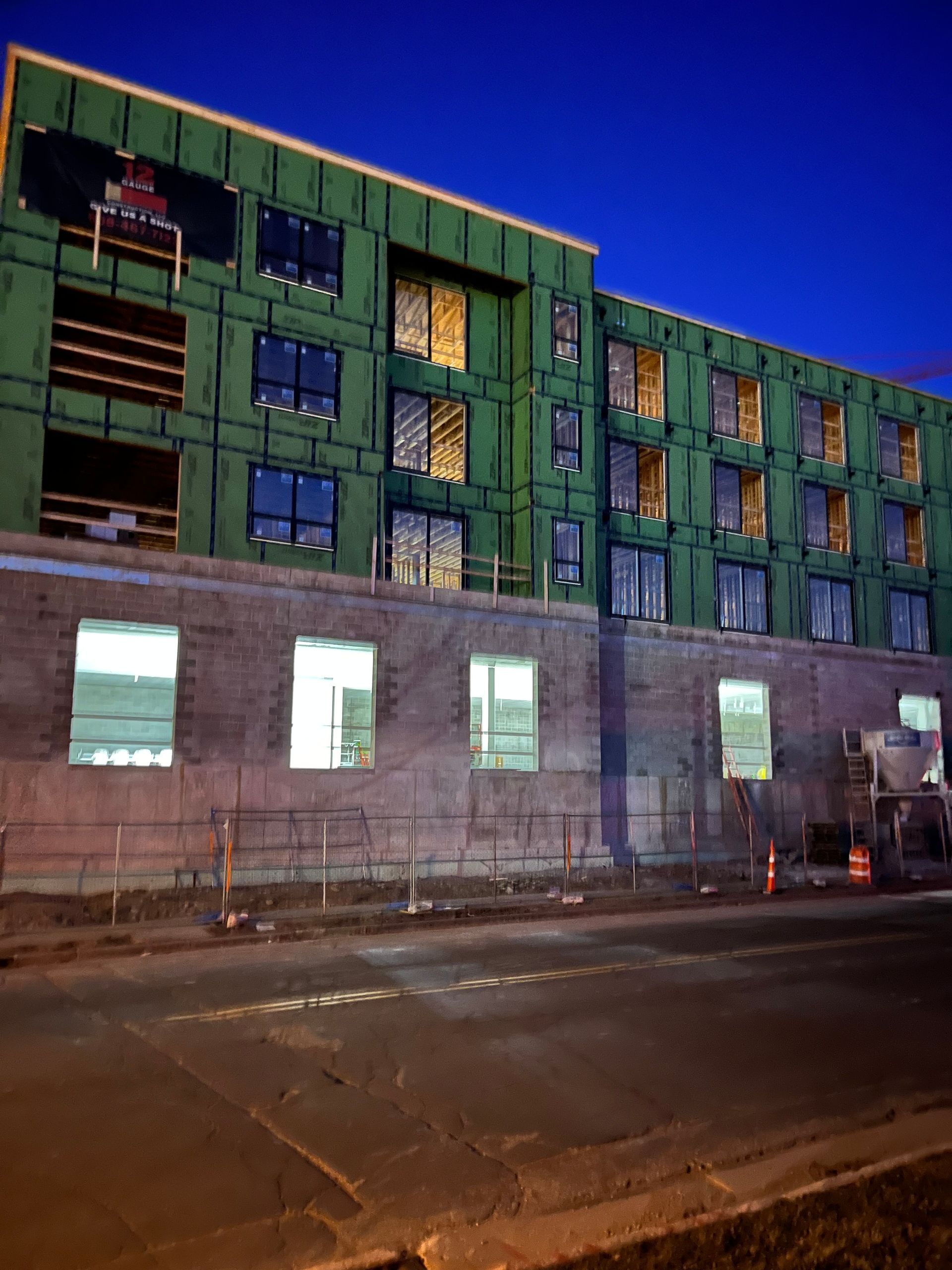 Building under construction at dusk; green siding, lit windows, concrete base.