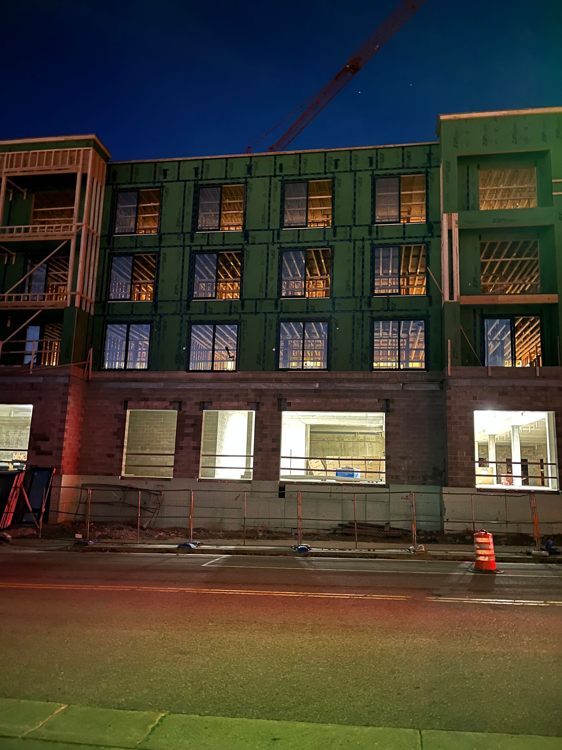 Building under construction at night, dark blue sky, exposed framing, lit windows, brick base, orange traffic cone.