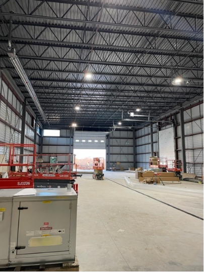 Inside of a large warehouse; open doorway, metal beam ceiling, concrete floor, equipment, and construction materials.