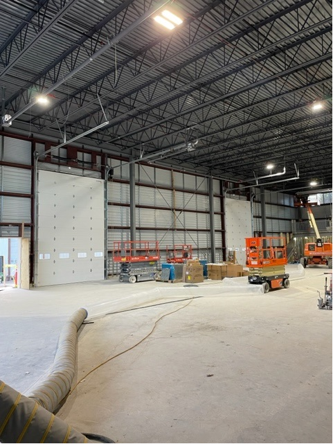 Interior view of a large warehouse under construction. White walls, exposed metal beams, equipment, and orange lifts.