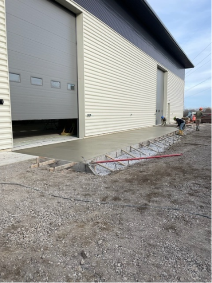 Exterior of a building with an open overhead door; a newly poured concrete ramp with workers.
