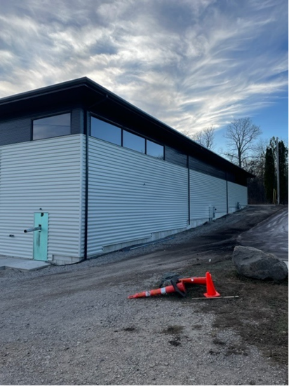 Side view of a light blue corrugated metal building with black trim, cloudy sky, orange traffic cone in foreground.