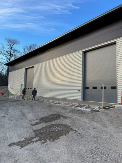 Exterior of a modern building with two large garage doors, two people stand near the doors on a gravel lot.