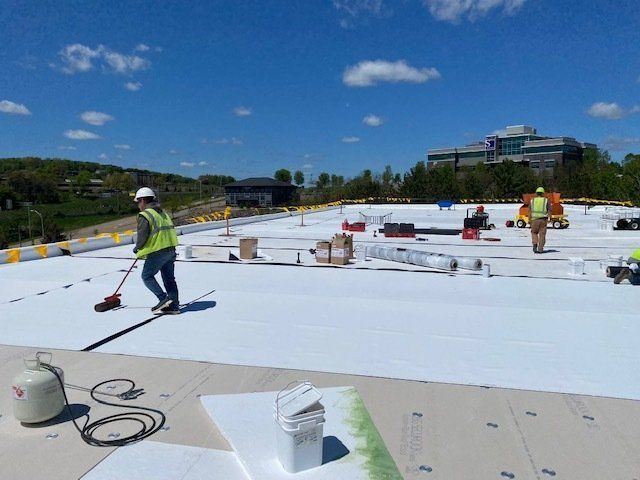 Workers installing white roofing material on a flat rooftop on a sunny day.