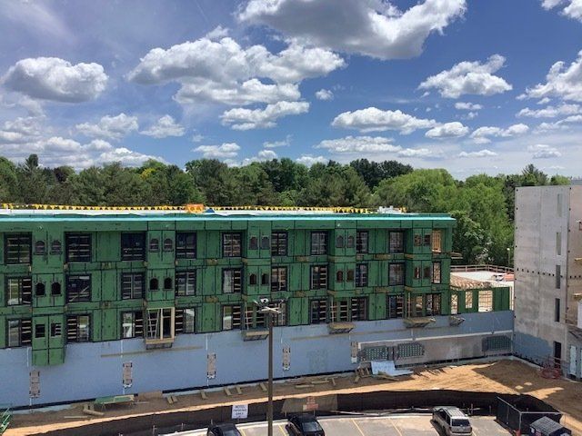 Building under construction with green siding, surrounded by trees and a parking lot. Blue sky with clouds overhead.