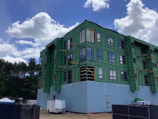 Building exterior under construction with green wrap and windows against a cloudy sky.