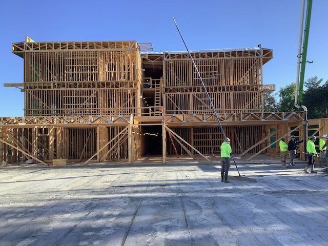 Wood-framed building under construction, workers in neon vests, concrete pump pouring concrete. Outdoors, sunny.