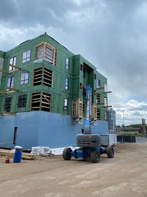 Building under construction; blue lift, green siding, brown wood framing, cloudy sky.