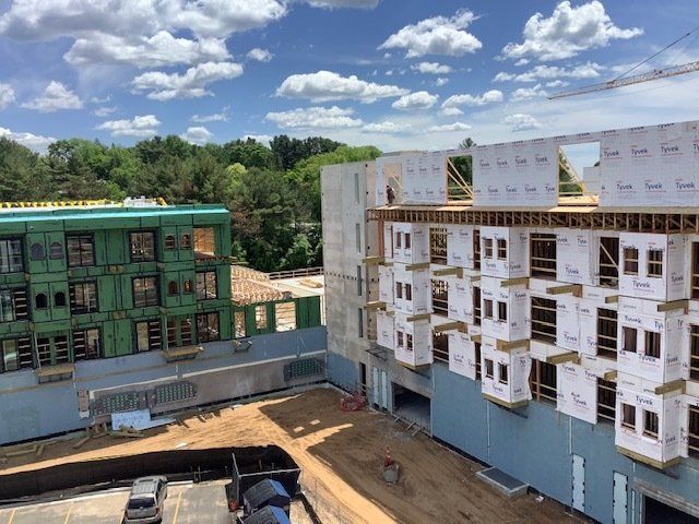 Two buildings under construction; one green-sheathed, one with sheathed window frames. Blue sky with clouds overhead.