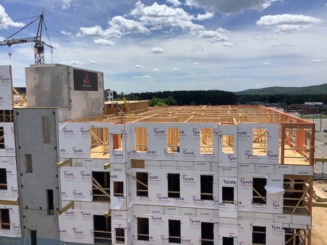 Construction site: multi-story building frame with wood beams, Tyvek wrap, under cloudy sky.