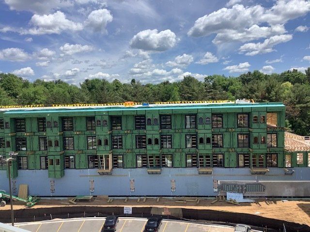 Building under construction, green sheathing, windows, blue sky, trees in background.