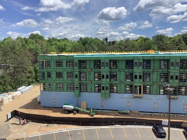 Construction site: multi-story building, wood framing, green weather barrier, surrounded by trees and dirt.