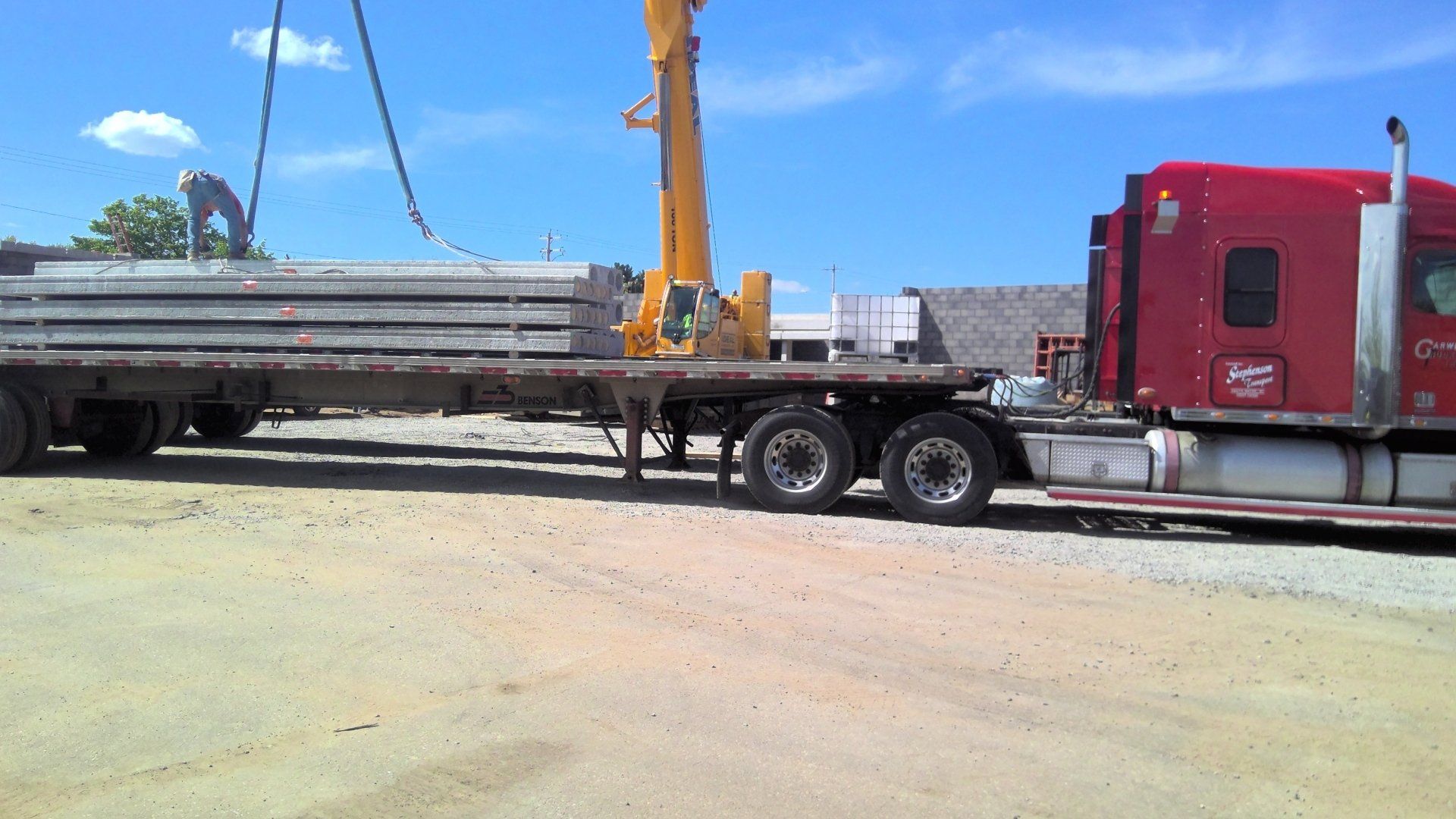 A crane lifting concrete beams off a flatbed truck on a sunny day.