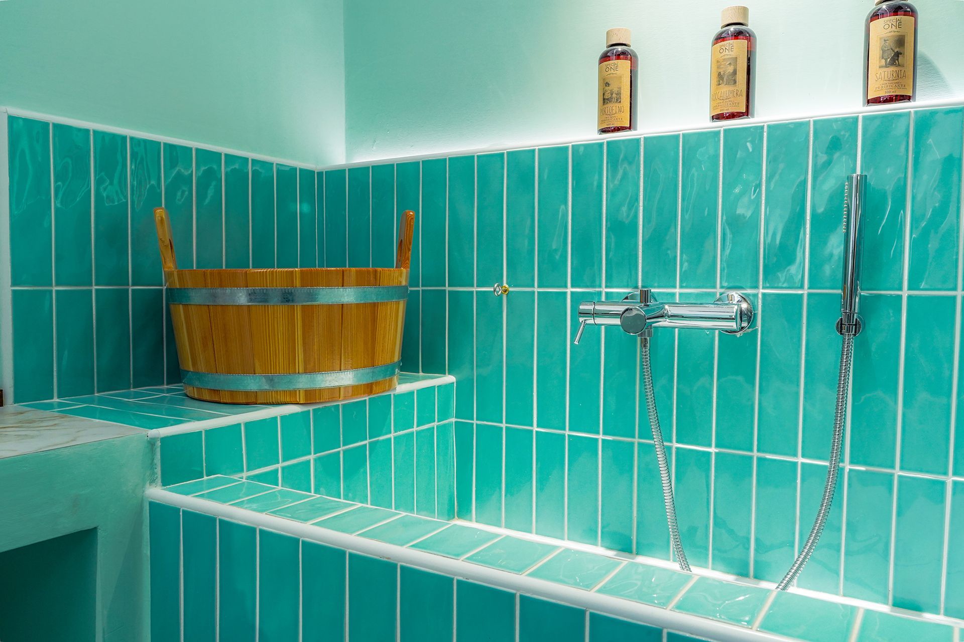Bagno con piastrelle turchesi, vasca da bagno, rubinetto cromato, portaoggetti da bagno in legno e flaconi sullo scaffale.