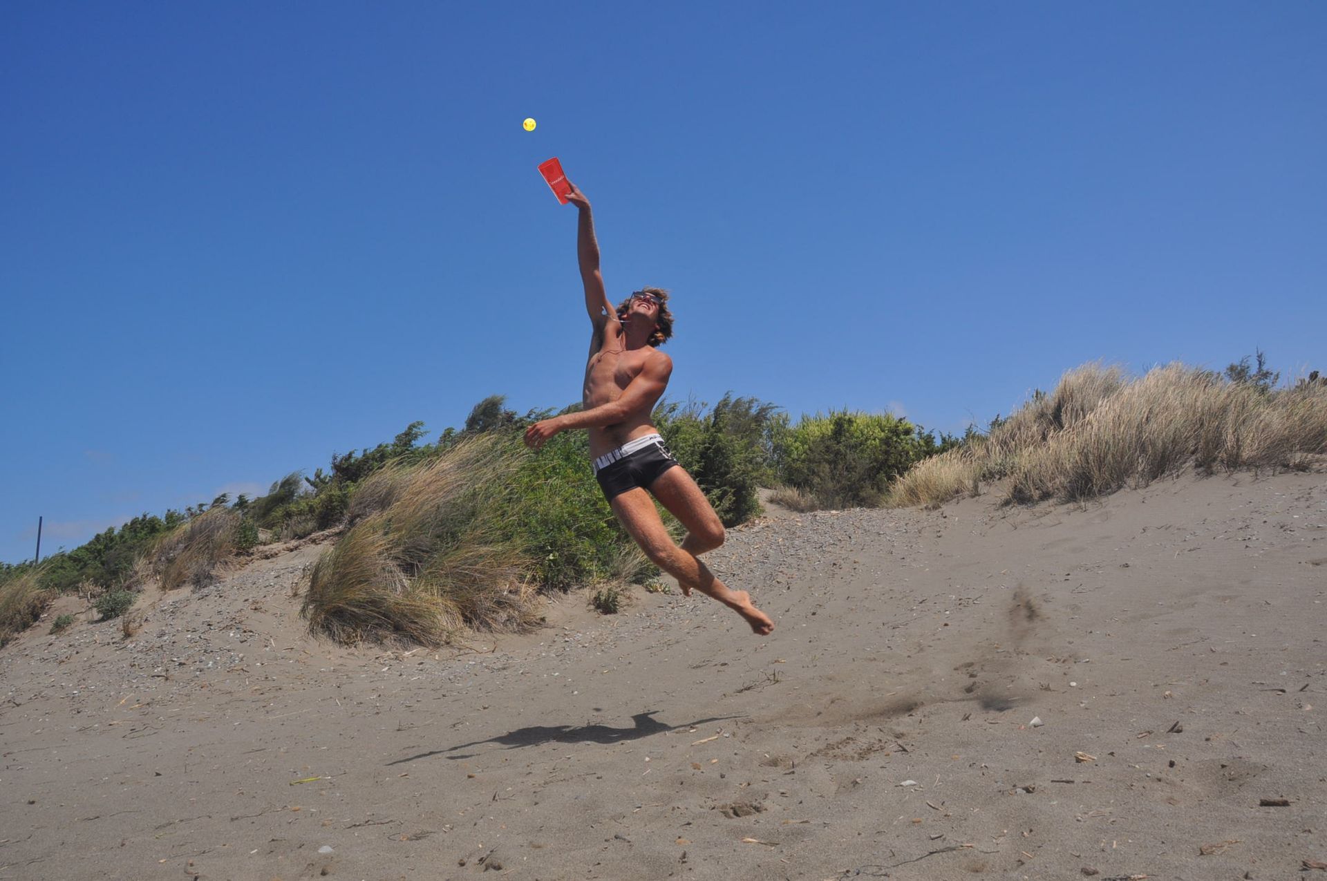Man leaping for a ball on a sandy beach, holding a red paddle. Blue sky, vegetation in the background.