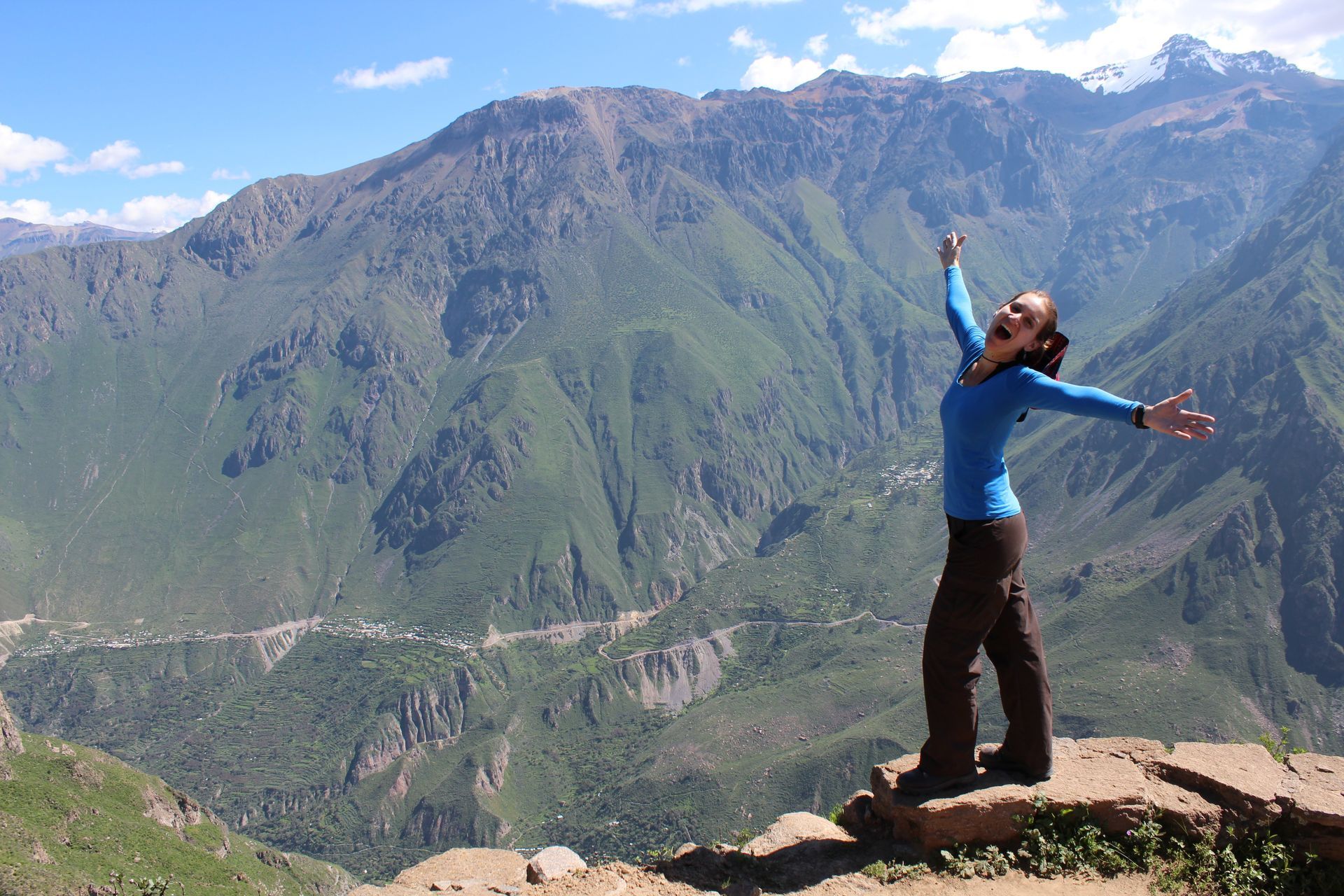 Woman with arms raised, smiles on a mountaintop, with expansive green valley and mountain range backdrop.