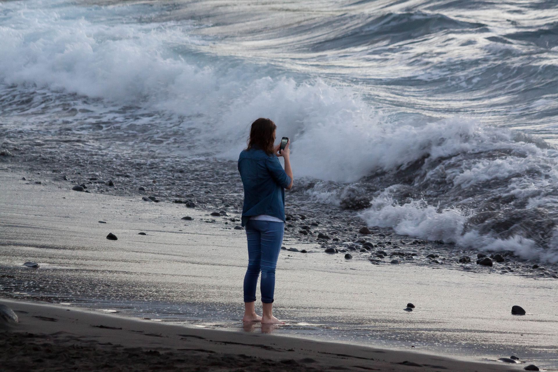 Woman taking a photo of ocean waves on a rocky beach. She wears a denim jacket and stands barefoot.