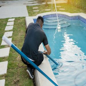 A person in a black shirt kneels by a pool, cleaning the water with a long blue pole.