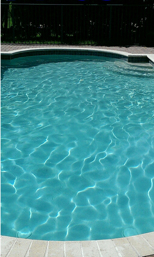 A clear, turquoise swimming pool with steps, bordered by stone coping and a dark fence in the background.