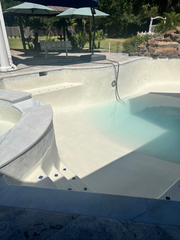 A partially filled swimming pool with light-colored plaster, built-in stairs, and a surrounding stone patio area.