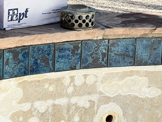 A pool edge featuring a row of mottled blue square tiles, a stone coping top, and a perforated circular drain cover.